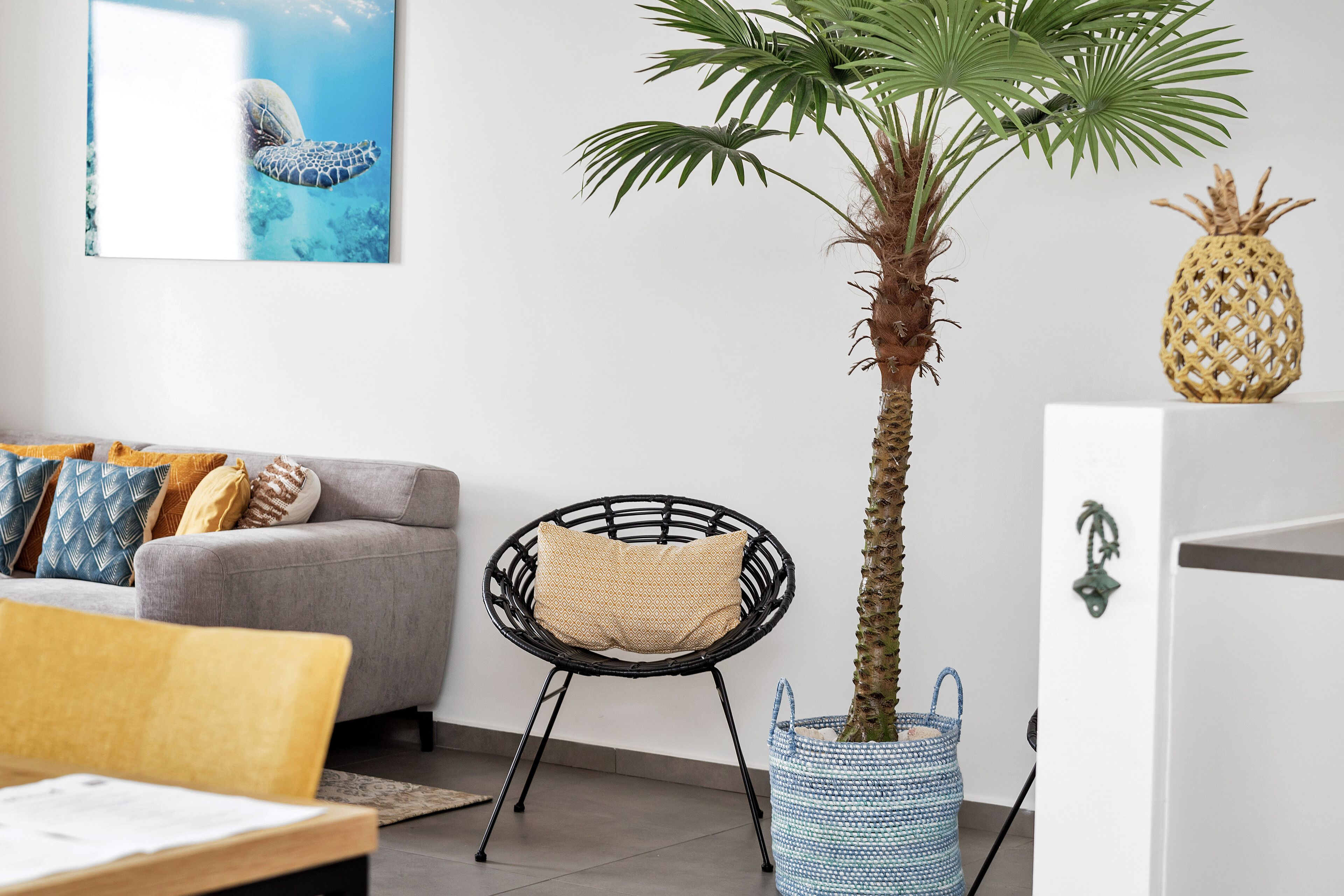 A living room with a grey sofa, a wicker chair, a potted palm tree, a woven basket, and a pineapple decoration.