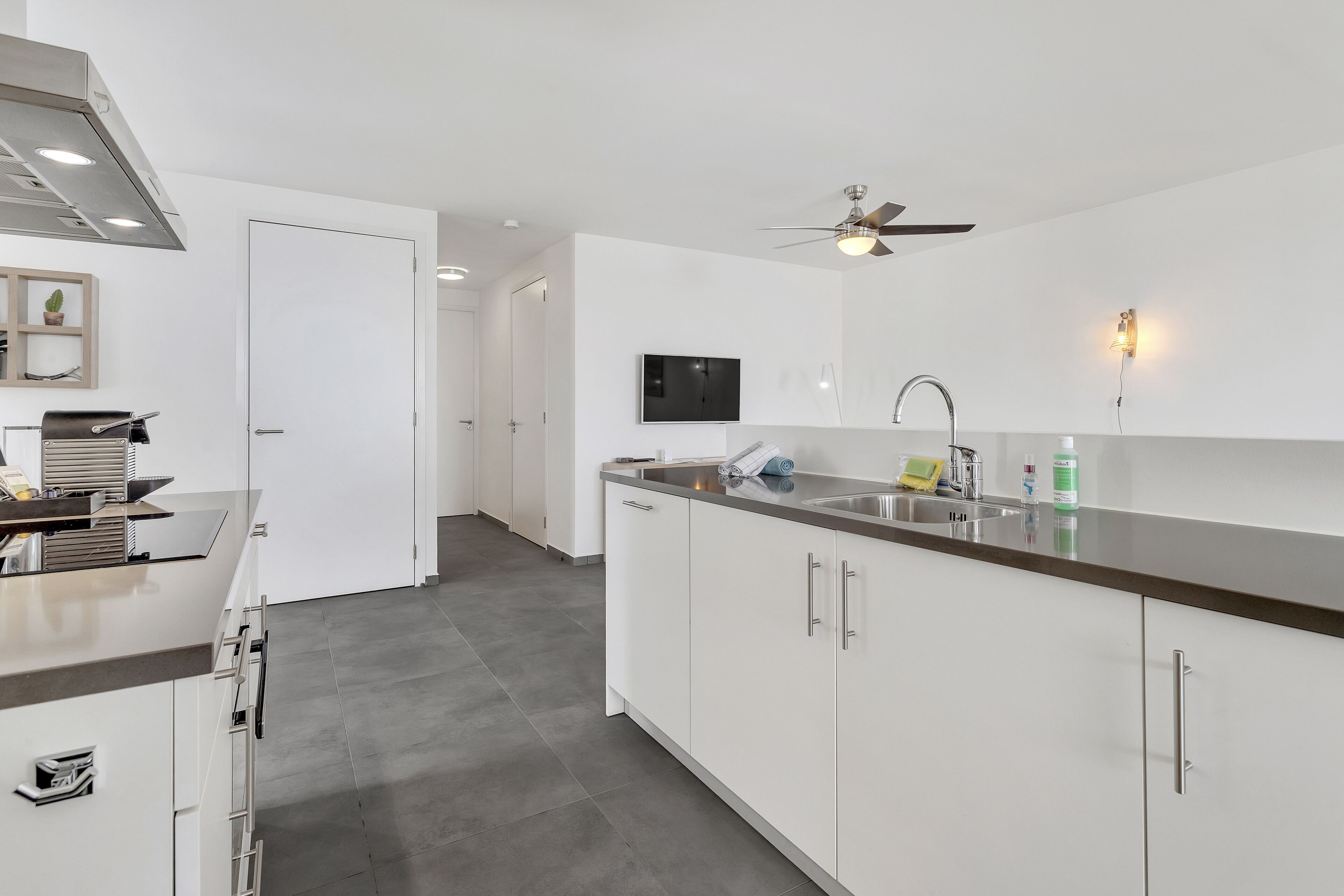 A modern kitchen with white cabinets, a stainless steel countertop, a built-in oven, and a ceiling fan.