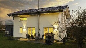 Ein modernes, zweistöckiges Haus mit Ziegeldach, Schornstein und einer Terrasse mit Gartenmöbeln.