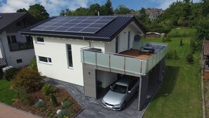 Ein modernes, zweistöckiges Haus mit Solardach, überdachter Auffahrt und einem kleinen Garten.