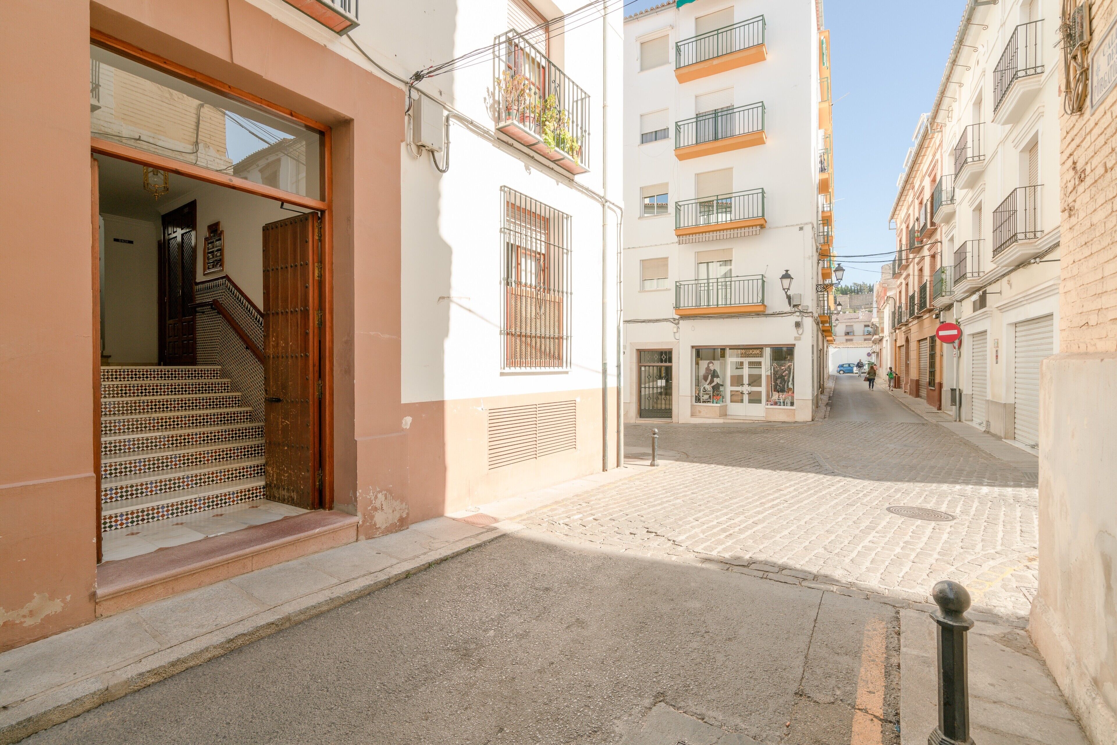 Una calle estrecha flanqueada por edificios, con una entrada visible y una escalera, bajo un cielo despejado.