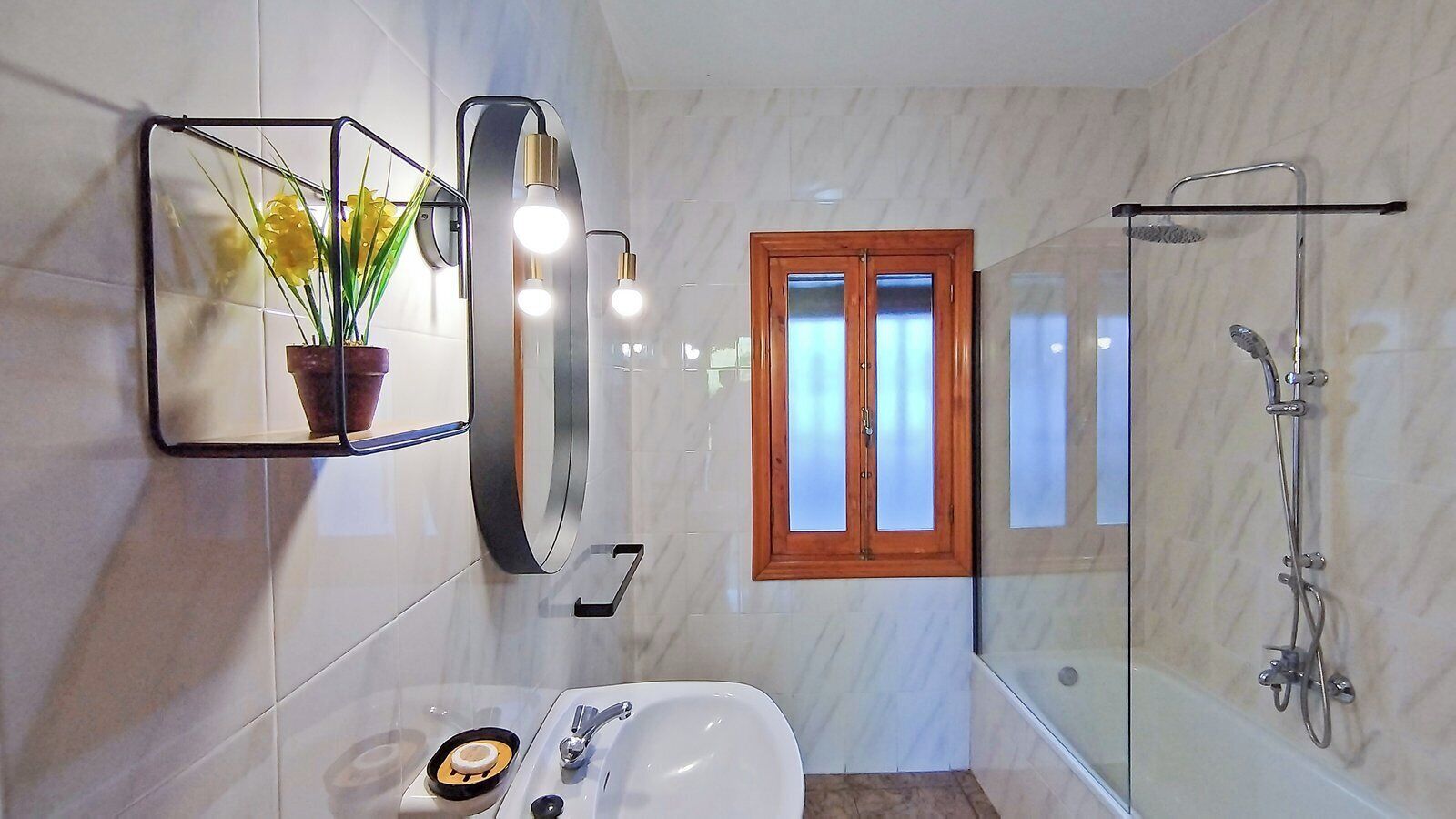 Un baño con cabina de ducha de vidrio, un lavamanos blanco, un espejo con planta en maceta y una ventana con vidrio esmerilado.