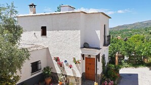 Un bâtiment blanc avec une porte en bois et des volets aux fenêtres, entouré de verdure et sous un ciel dégagé.