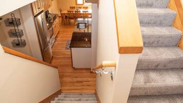 Un escalier mène à une cuisine dotée d’un plancher en bois, d’un réfrigérateur en acier inoxydable et d’un coin repas avec des chaises et une table en bois.