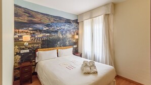 Une chambre d’hôtel avec un grand lit, une table de chevet, une lampe et une fresque représentant un paysage montagneux.