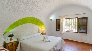 Ein Schlafzimmer mit gewölbter weißer Decke, einem Bett mit weißem Überwurf, einem Nachttisch mit Lampe und einem Fenster mit Vorhängen.