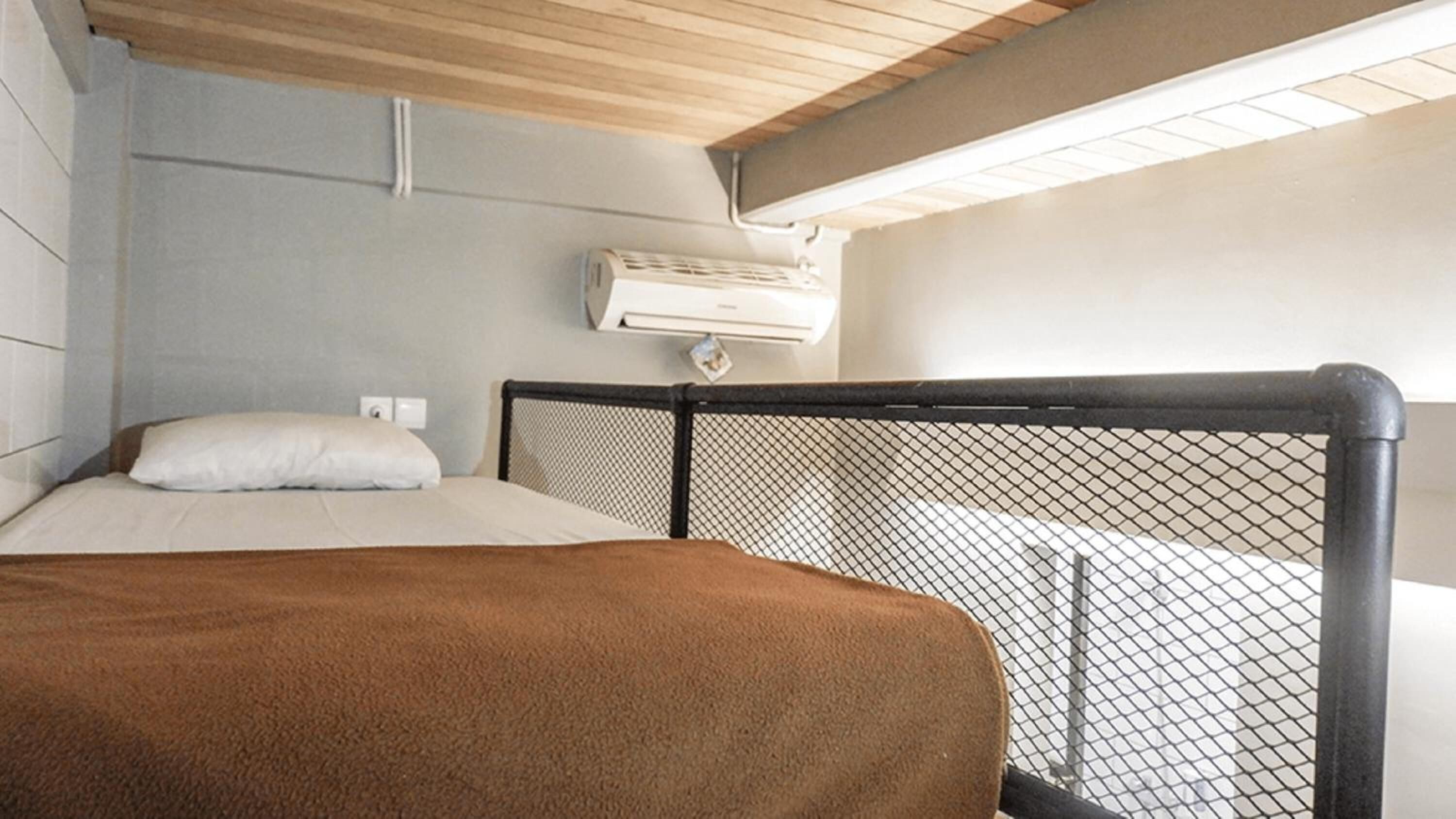 A bedroom with a bed, a headboard with a mesh design, an air conditioning unit on the wall, and a wooden ceiling.