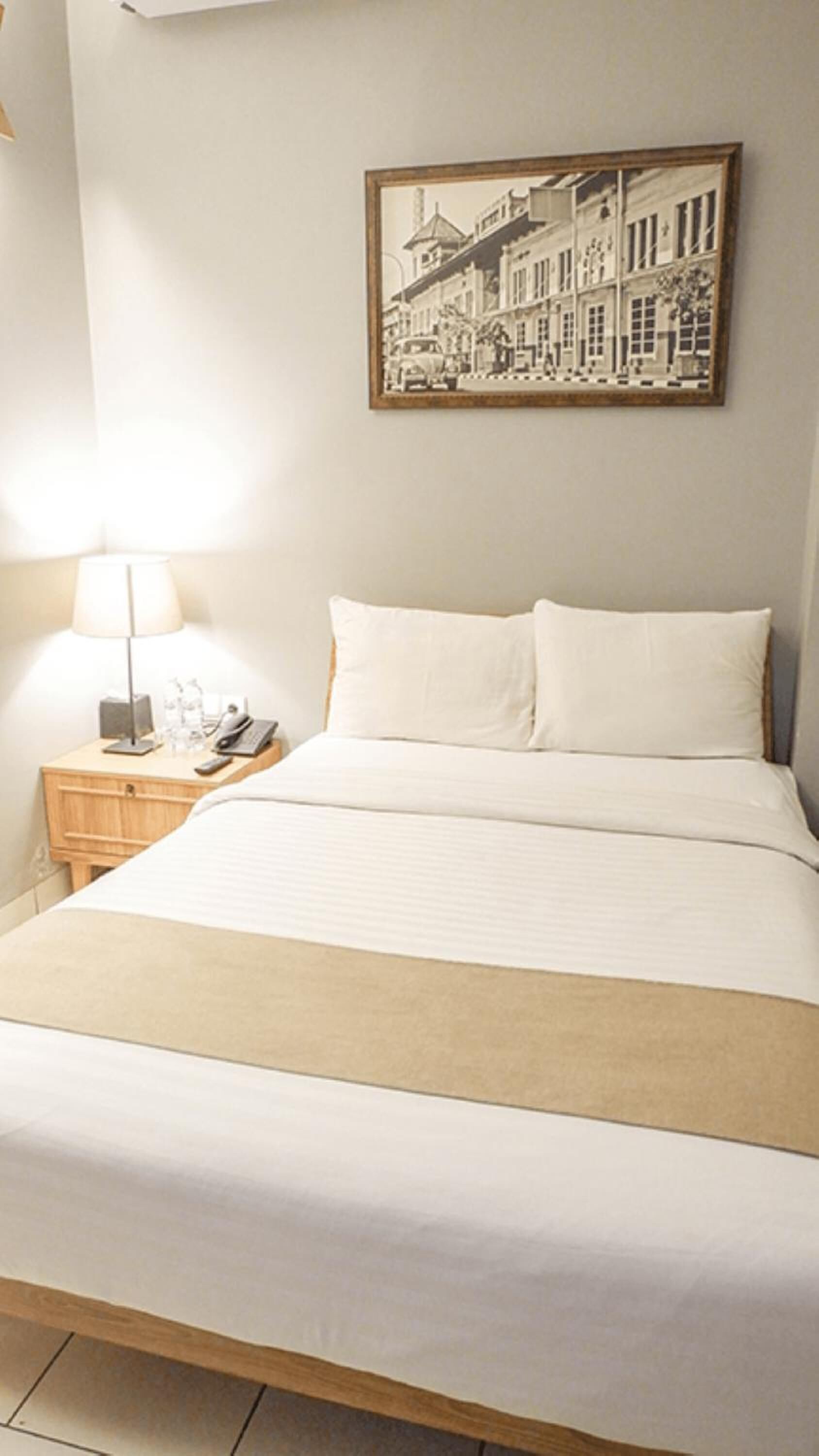A hotel room with a bed, a nightstand with a lamp, a phone, and a framed picture on the wall.