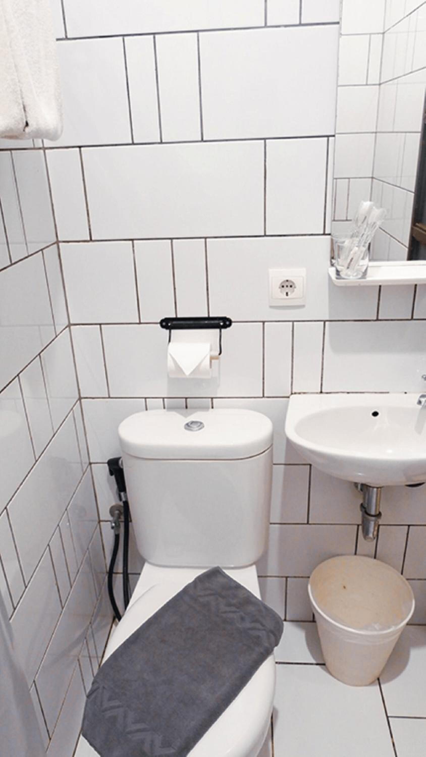 A small, clean bathroom with white tiles, a toilet, a sink, and a trash bin.