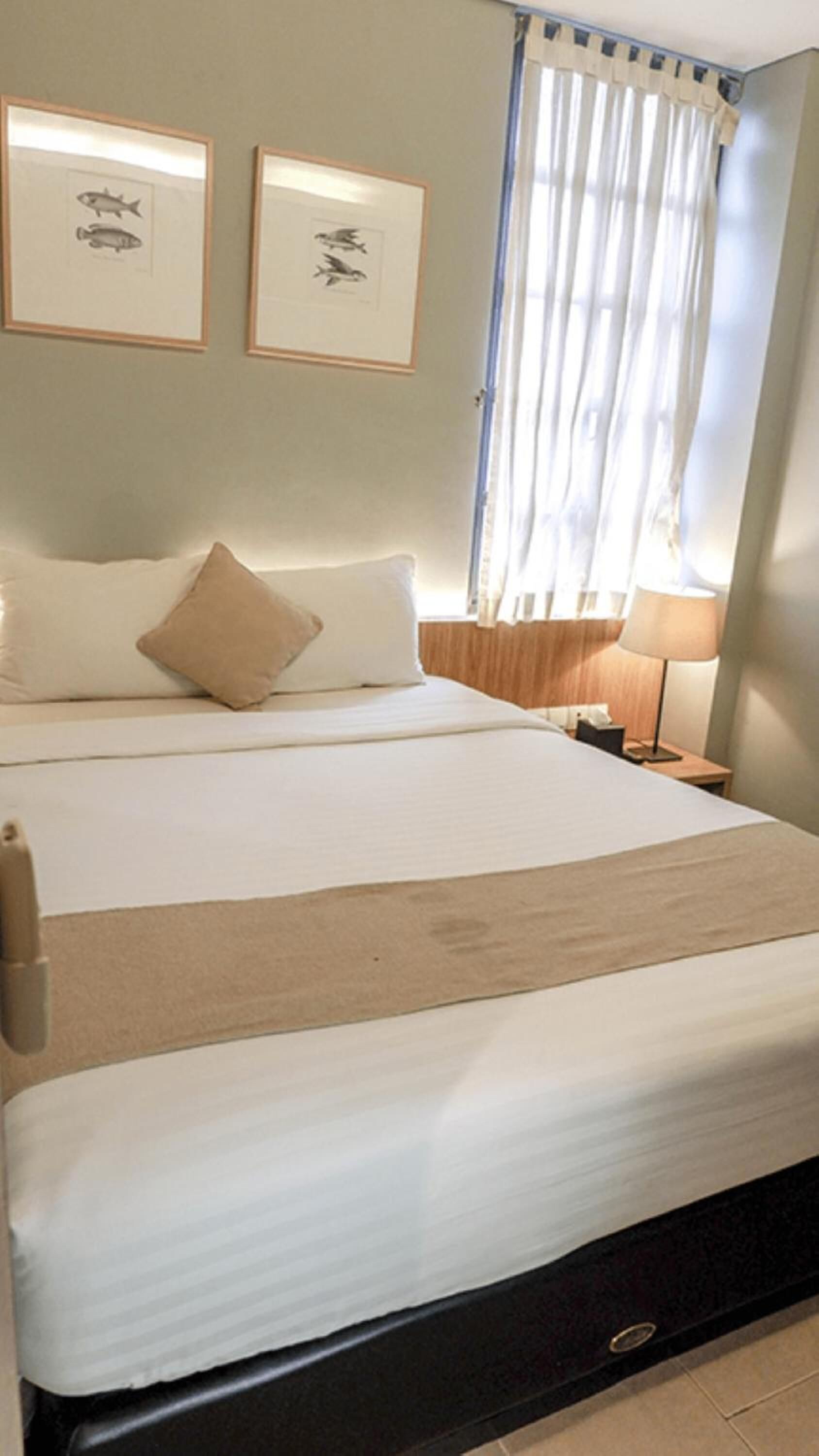 A hotel room with a large bed, two framed pictures on the wall, a window with curtains, and a bedside table with a lamp.