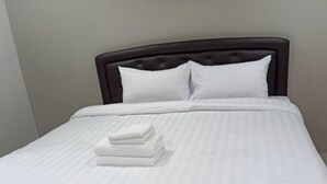 Una cama bien hecha con sábanas y almohadas blancas, un cabecero oscuro y una pequeña pila de toallas dobladas al pie.