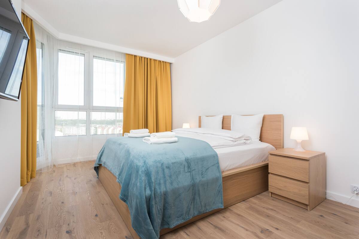 A modern hotel room with a bed, bedside table, and a large window with curtains.