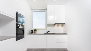 A modern kitchen with white cabinetry, a built-in oven, a window above the sink, and a grey floor. - Apartamenty InPoint Mogilska Tower (Kraków)