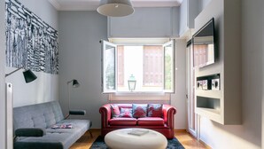 A modern living room with a grey sofa, a red sofa with patterned cushions, a white ottoman, a black rug, and a large window with shutters. - The Burton Luxury 1BR in Plaka, Athens (Athina)