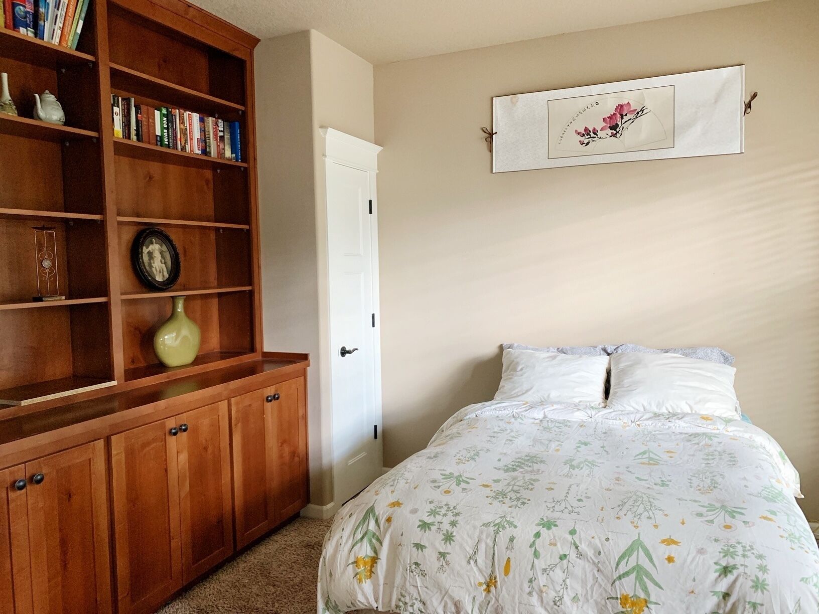A bedroom with a wooden bookshelf, a bed with floral bedding, and a framed artwork on the wall.