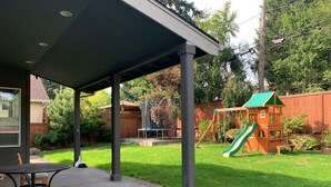 En overdækket patio med bord og stole, en legeplads i træ med rutsjebane, og en lukket gårdhave med træer og et træhegn.