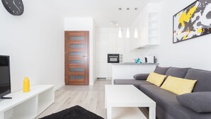 Una sala de estar moderna con un sofá gris, una mesa de centro blanca y una alfombra negra. Hay un televisor sobre un mueble blanco y una botella amarilla sobre el mueble. Una puerta de madera da a una cocina con armarios blancos y un microondas.