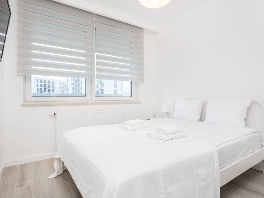 A clean, white bedroom with a bed, a radiator, a window with blinds, and a view of buildings outside.