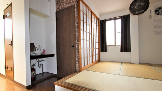 Une pièce de style japonais traditionnel, avec un sol recouvert de tatamis, une porte coulissante, un petit coin cuisine et une fenêtre avec des rideaux.