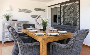 Un coin repas avec une table en bois dressée pour quatre personnes, entourée de chaises en osier, d’une plante en pot et d’une décoration murale représentant des poissons.