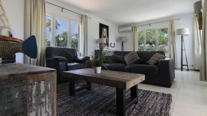 A living room with a sofa, a coffee table, and a lamp. There is a window with curtains and a view of palm trees.