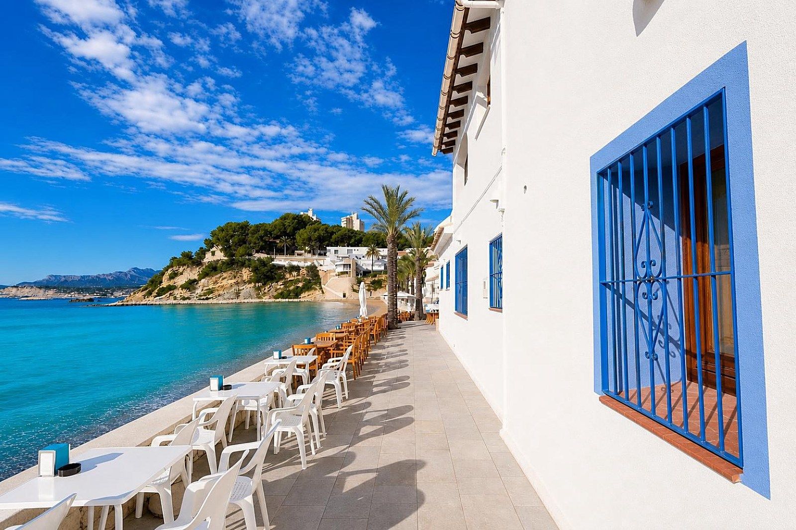 A seaside terrace with white tables and chairs, a blue railing, and a view of the sea and a hill with buildings.
