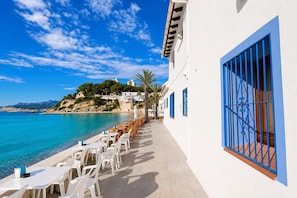 A seaside terrace with white tables and chairs, a blue railing, and a view of the sea and a hill with buildings.