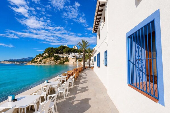 A seaside terrace with white tables and chairs, a blue railing, and a view of the sea and a hill with buildings.