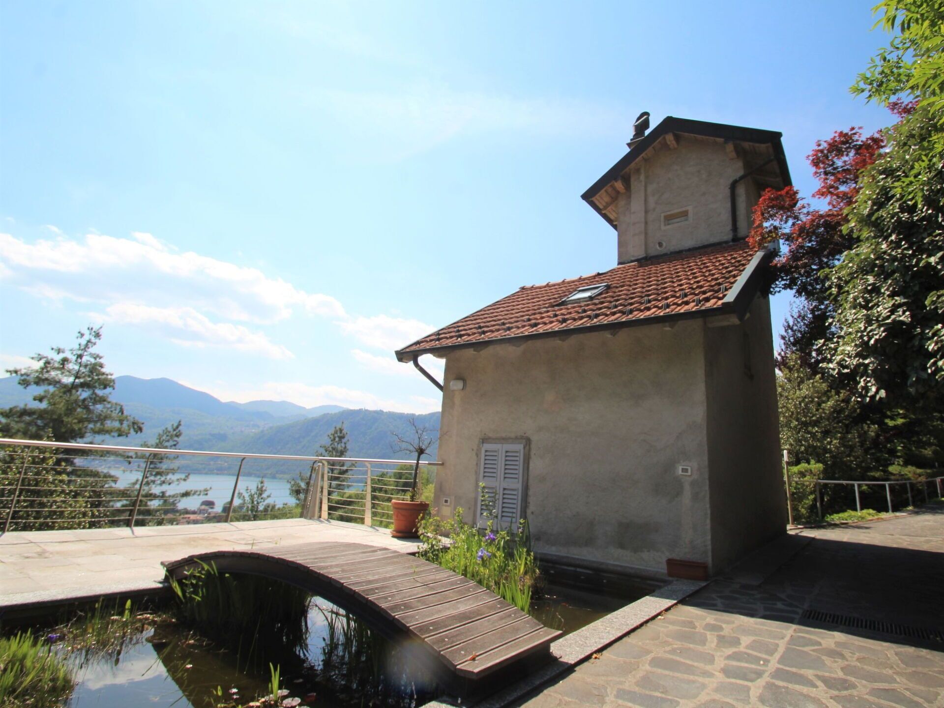 A small building with a tiled roof, a wooden bridge over a pond, and a view of mountains in the background.