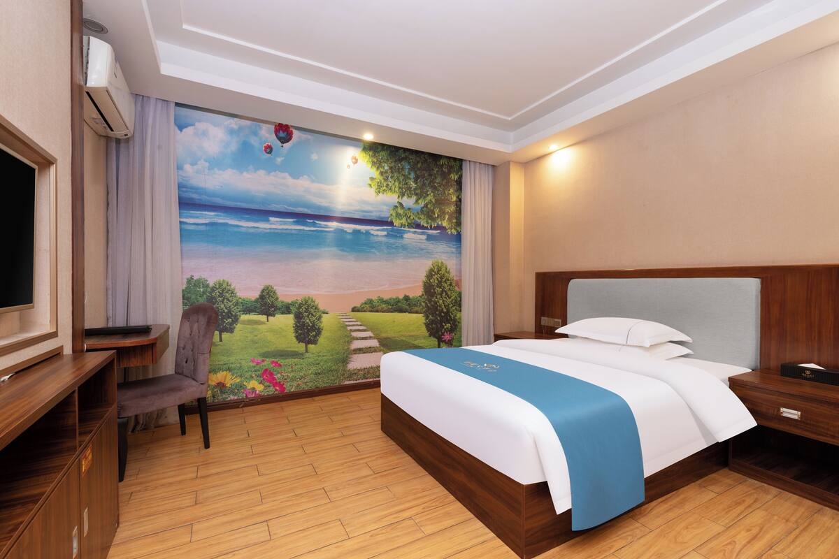 Een hotelkamer met een groot muurschildering van een strand, een bed met een blauwe sprei, een houten nachtkastje, een bureau met een stoel en een airconditioner.