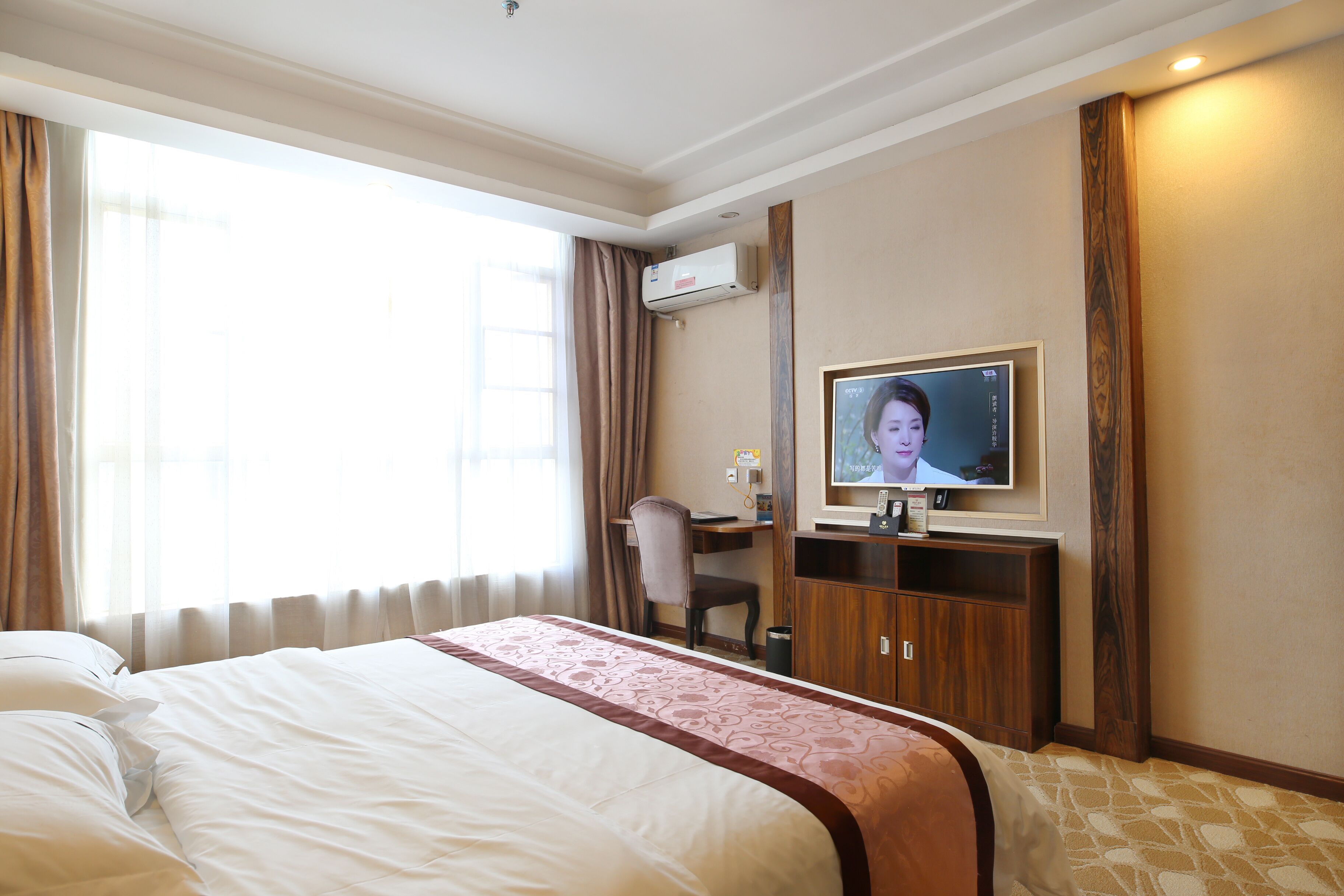 A hotel room with a large bed, a desk, a chair, an air conditioning unit, and a television.