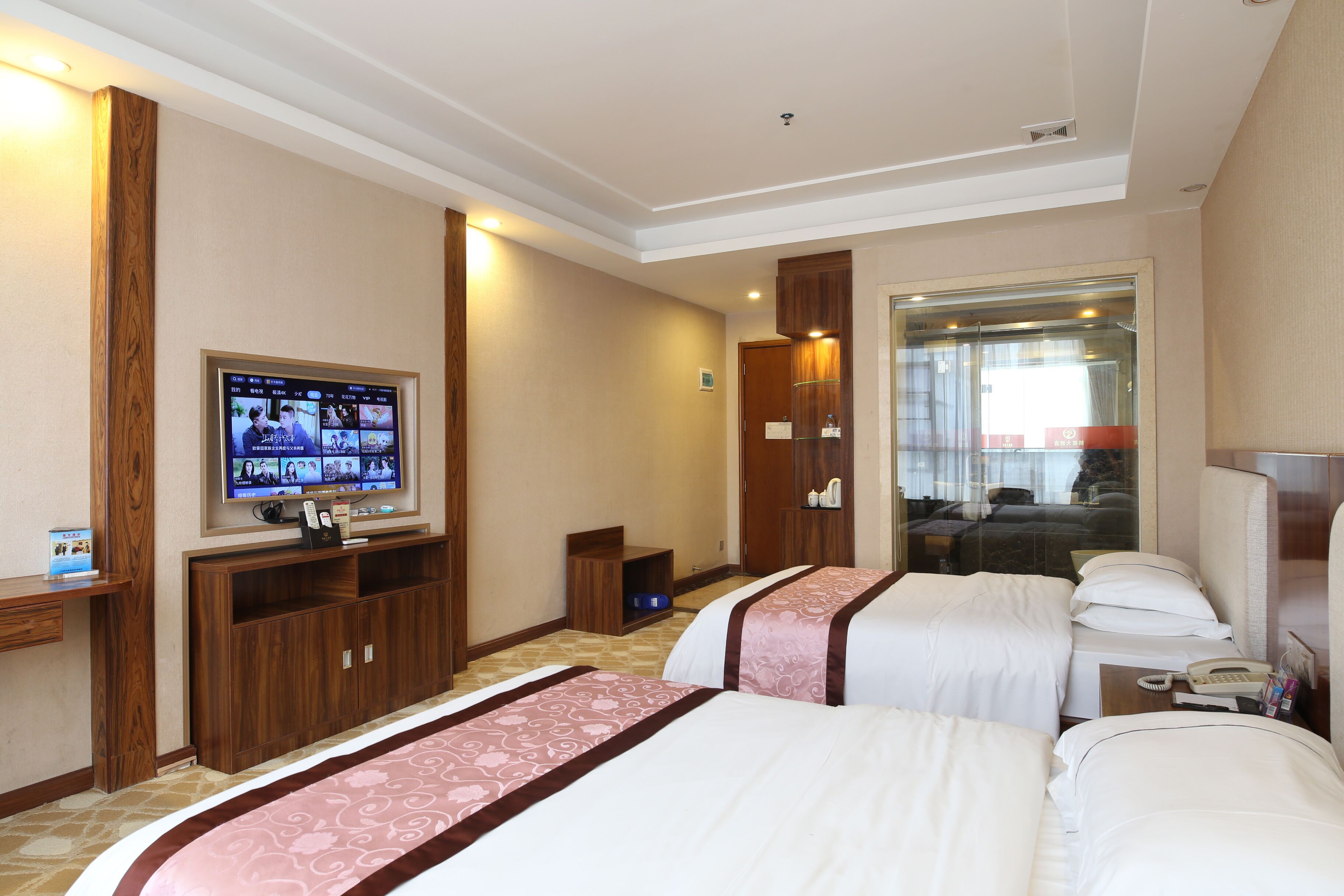 Quarto de hotel com duas camas, televisão, escrivaninha e banheiro visível através de uma porta de vidro.