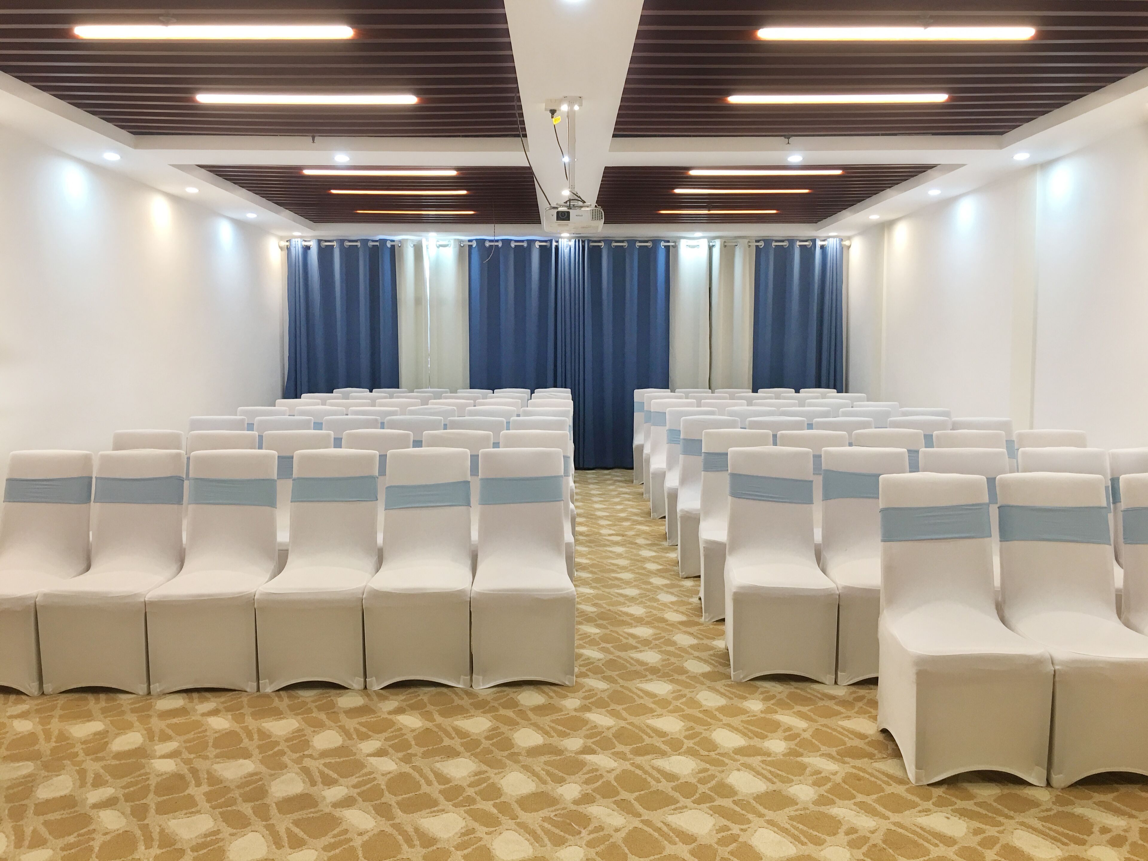 Un salón de conferencias con filas de sillas blancas, cortinas azules y una alfombra estampada.