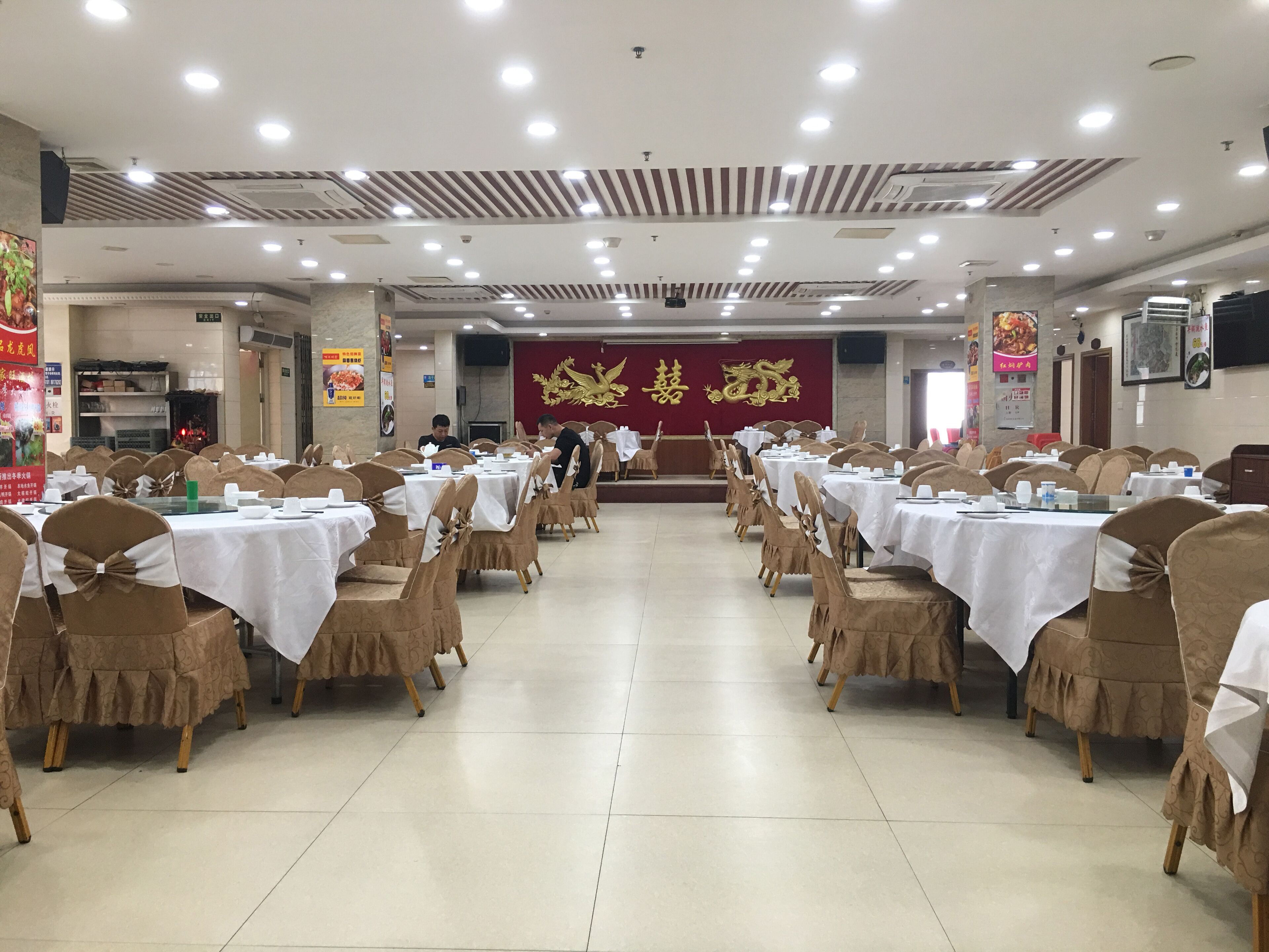 A banquet hall with tables set for a formal event, featuring a stage with a golden dragon emblem, and a tiled floor.