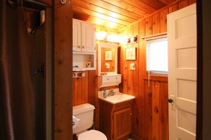 Compact bathroom with a toilet, sink, and mirror, featuring a shelf and window with blinds.