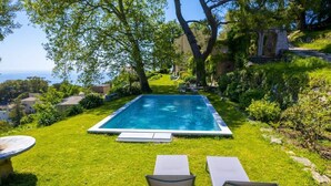 A rectangular swimming pool with a clear blue water surface, surrounded by well-maintained grass and trees. There are two lounge chairs and a small table on the grass. - Villa A Ventosa (Bastia)