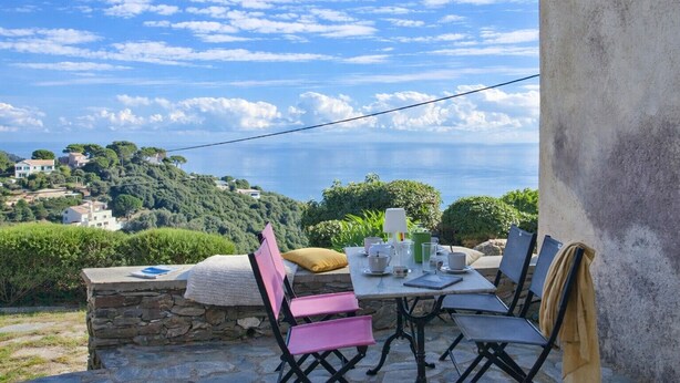 An outdoor dining area with a table set for two, overlooking a coastal landscape with houses and greenery. - Villa A Ventosa (Bastia)