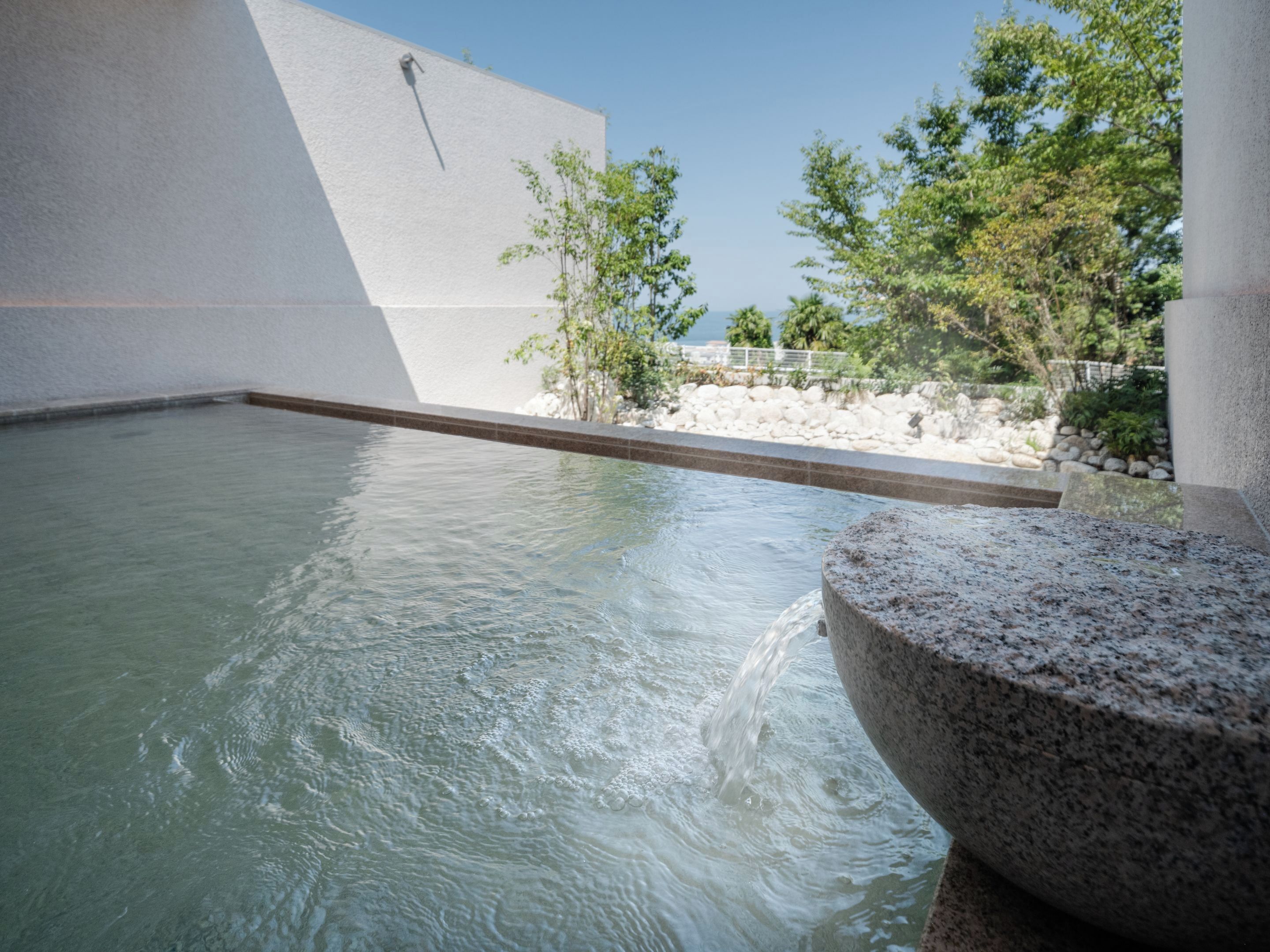 Un jacuzzi con agua cristalina, una cuenca de piedra y vistas a árboles y una pared blanca.