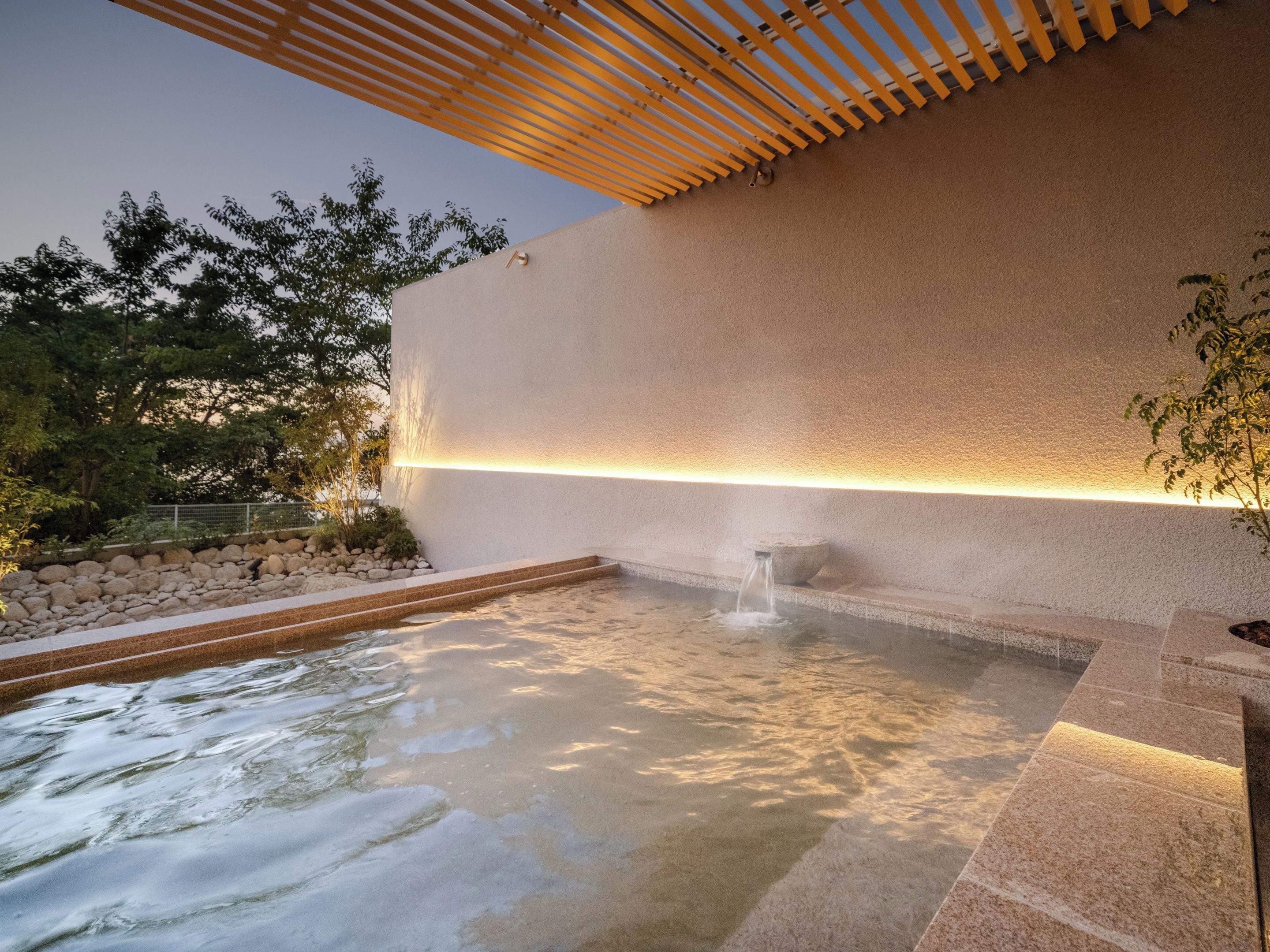 A spa area with a stone pool, a wooden pergola, and a wall with a light strip.