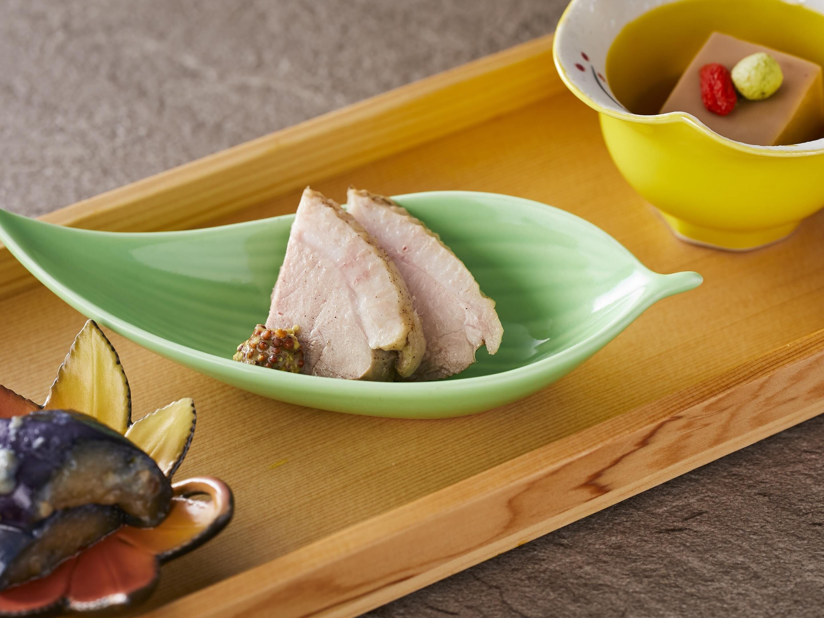 Una bandeja de madera con un plato en forma de hoja verde que contiene carne en rodajas y un recipiente pequeño con salsa, junto a un tazón amarillo con dos dulces redondos.