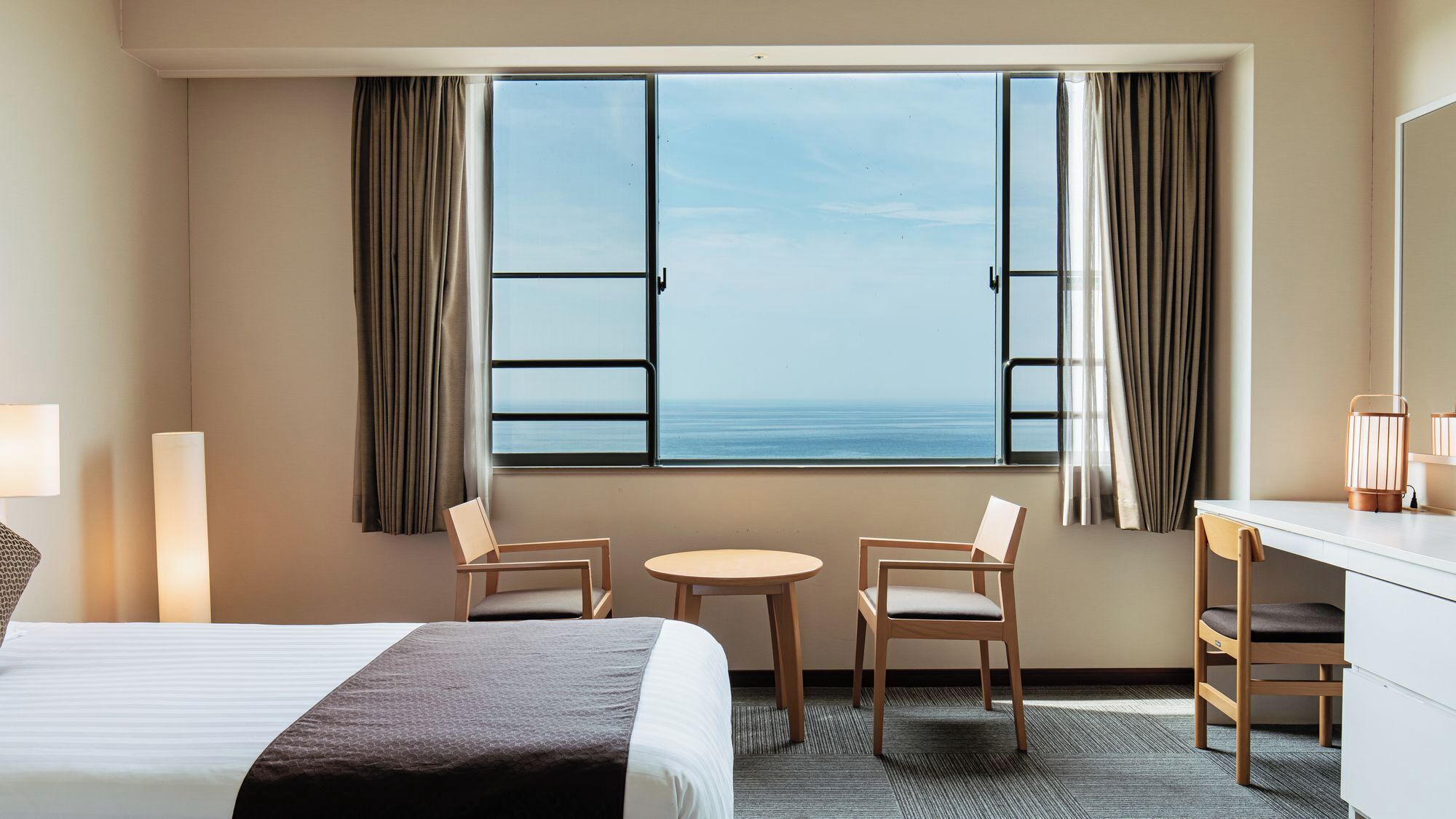A hotel room with a bed, two chairs, a small table, a desk, and a large window with curtains.
