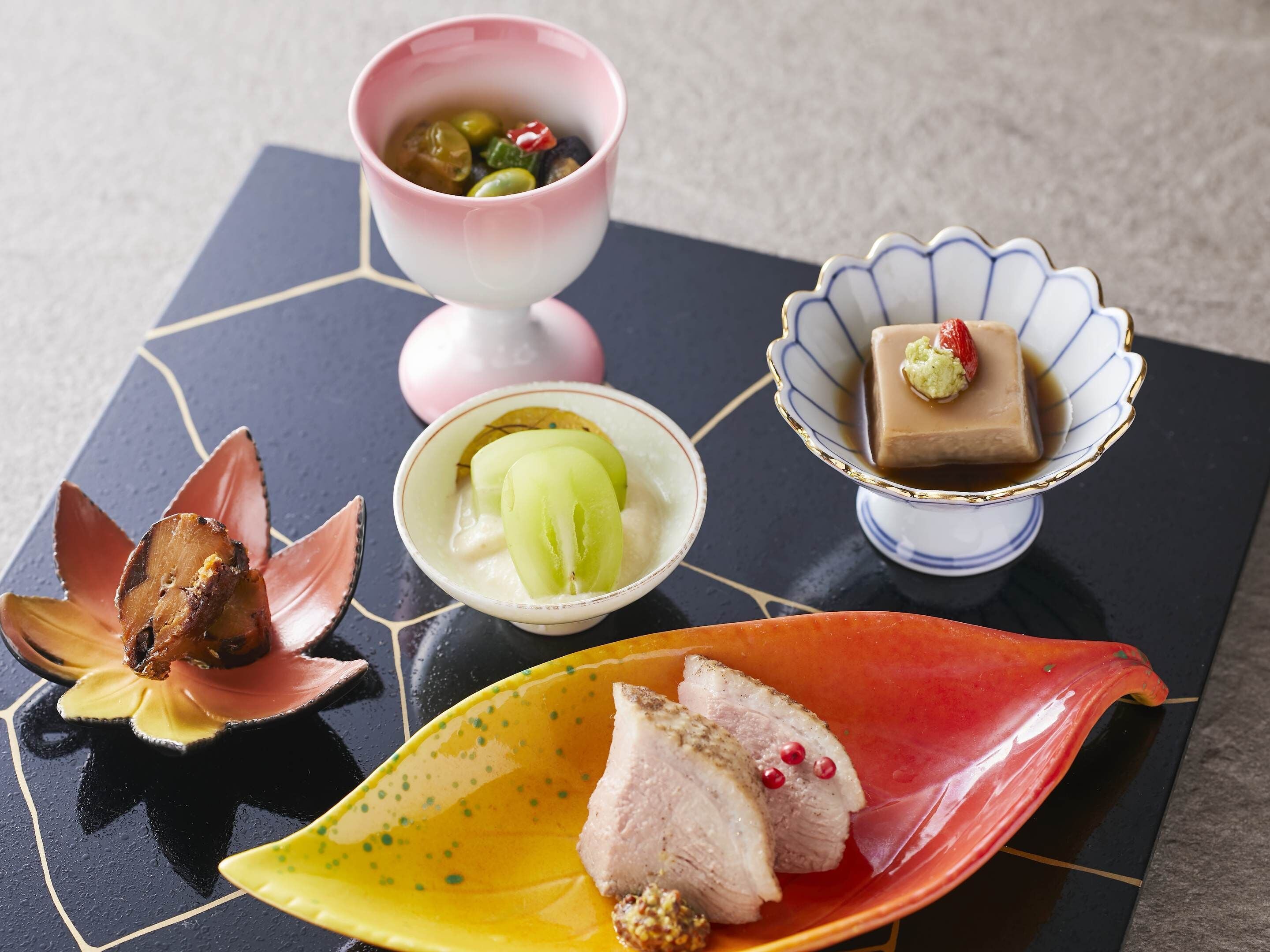 A variety of food items on a table, including a bowl of soup, a dessert in a scalloped dish, and a plate with sliced meat.