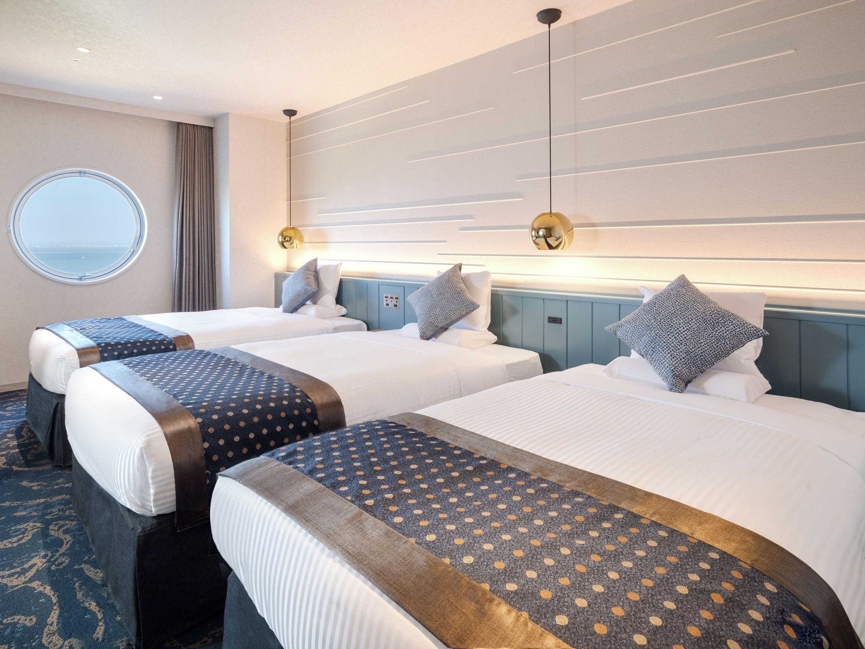 A hotel room with two beds, a round window, and a view of the sea.