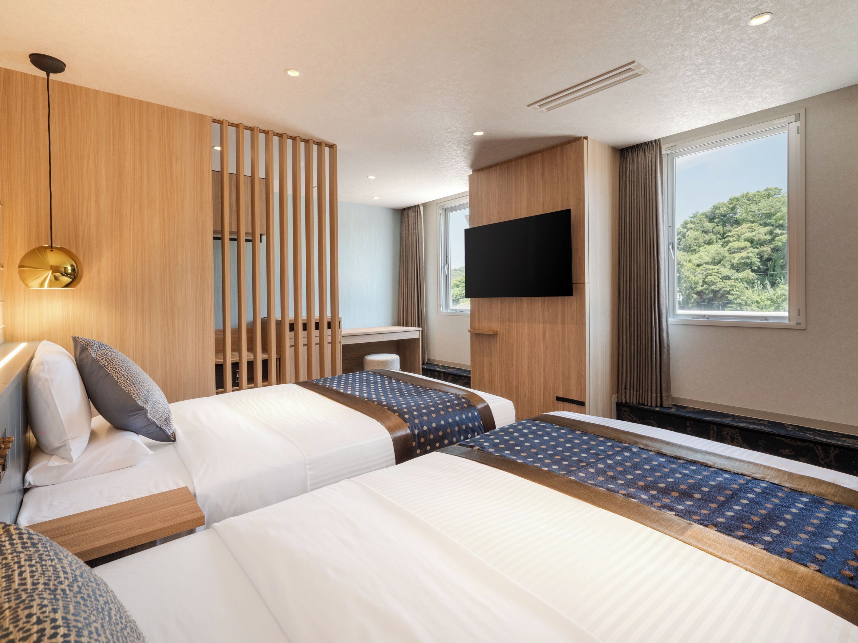 A hotel room with two beds, a desk, a TV, and a window with a view of greenery.