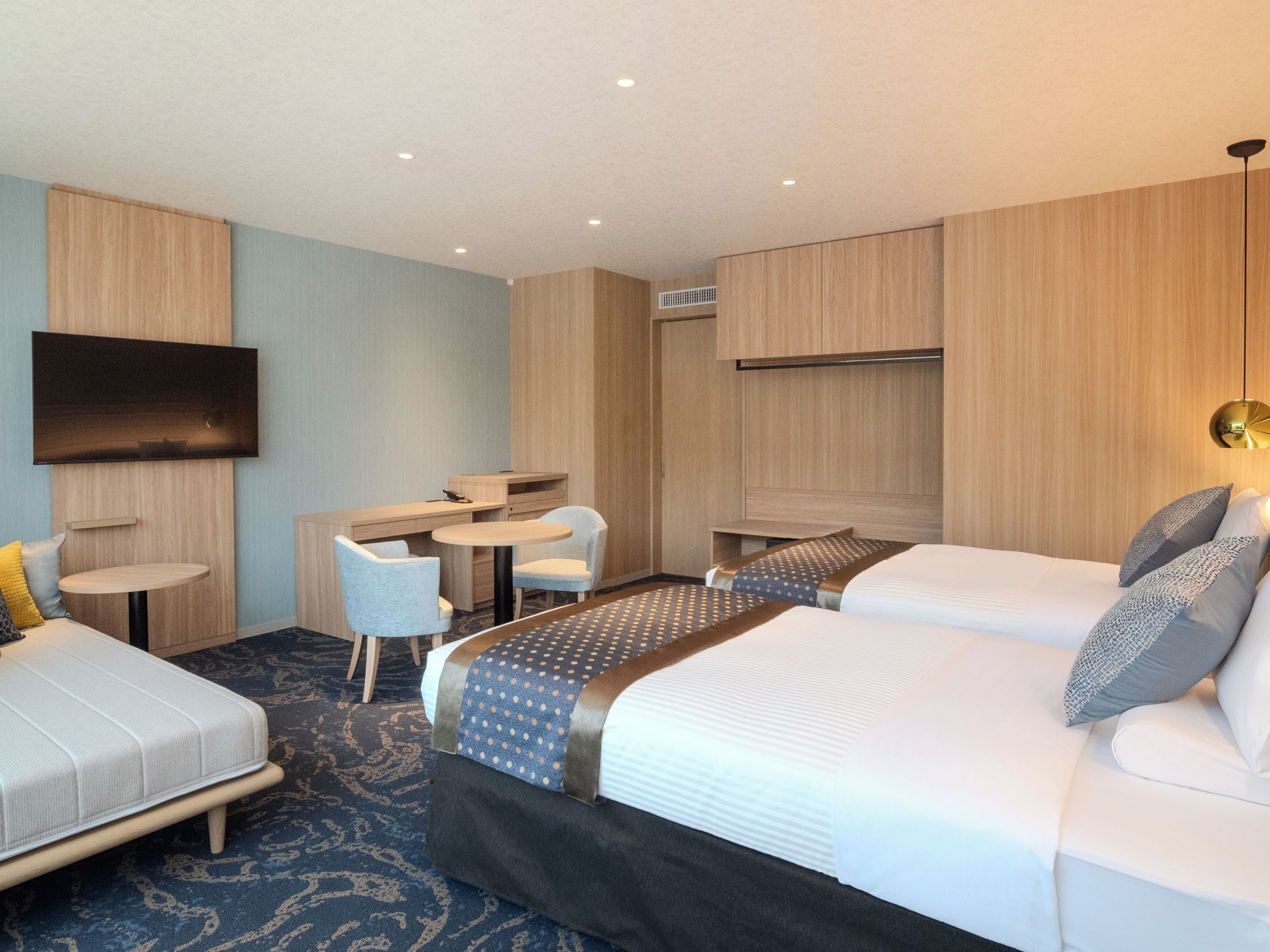 A hotel room with two beds, a desk, a chair, a TV, and a ceiling fan.