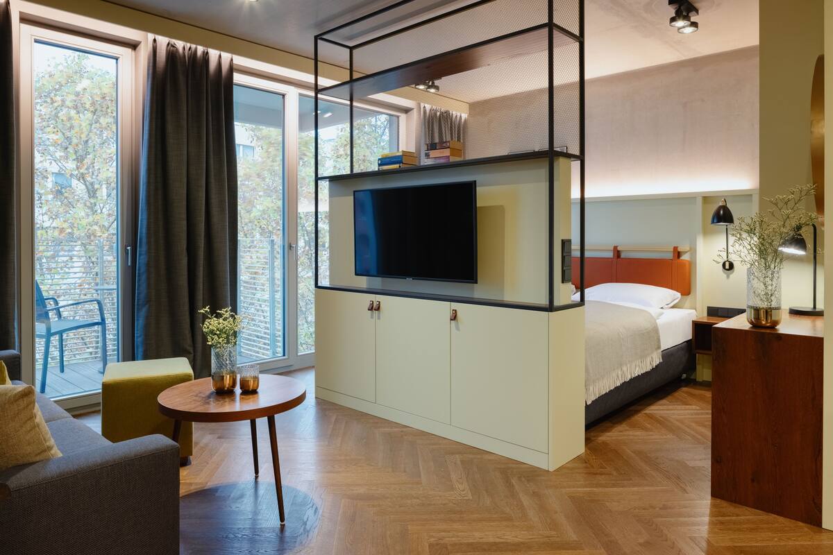 Pieni hotellihuone, jossa on sänky, seinälle asennettu televisio, pieni pöytä ja sohva.