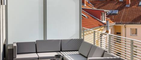 A balcony with a grey sectional sofa, a wicker coffee table, and a glass partition.
