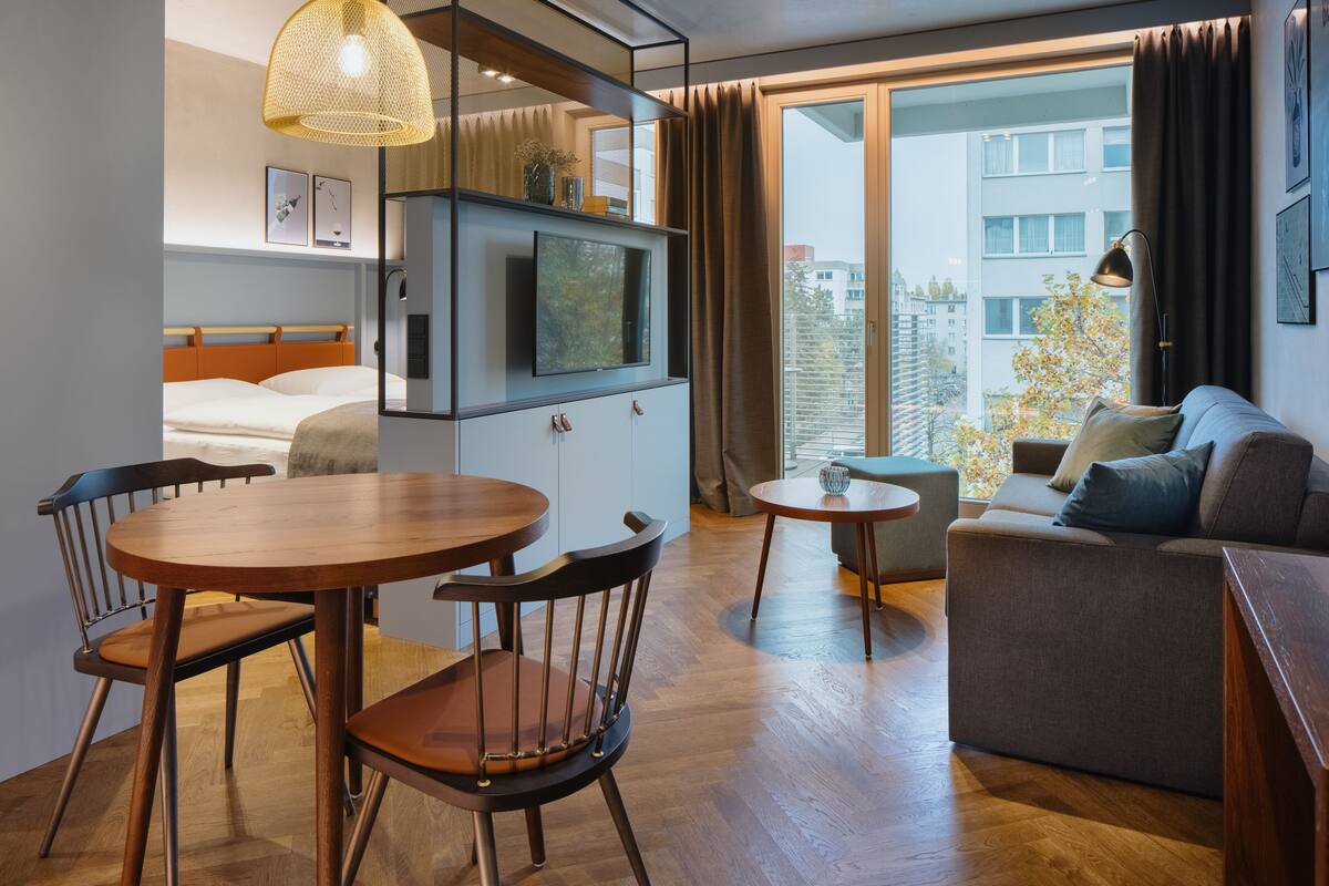 Moderni hotellihuone, jossa on puinen ruokapöytä, sohva, televisio ja suuri ikkuna, josta avautuu näkymä kaupunkiin.