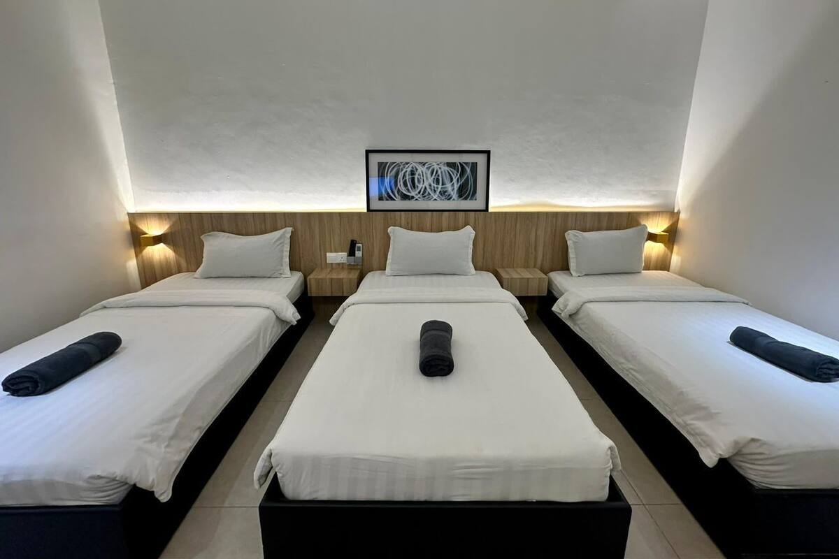 A hotel room with three single beds, each with a pillow and a rolled towel. There is a framed artwork above the beds and a small table with a phone on it.