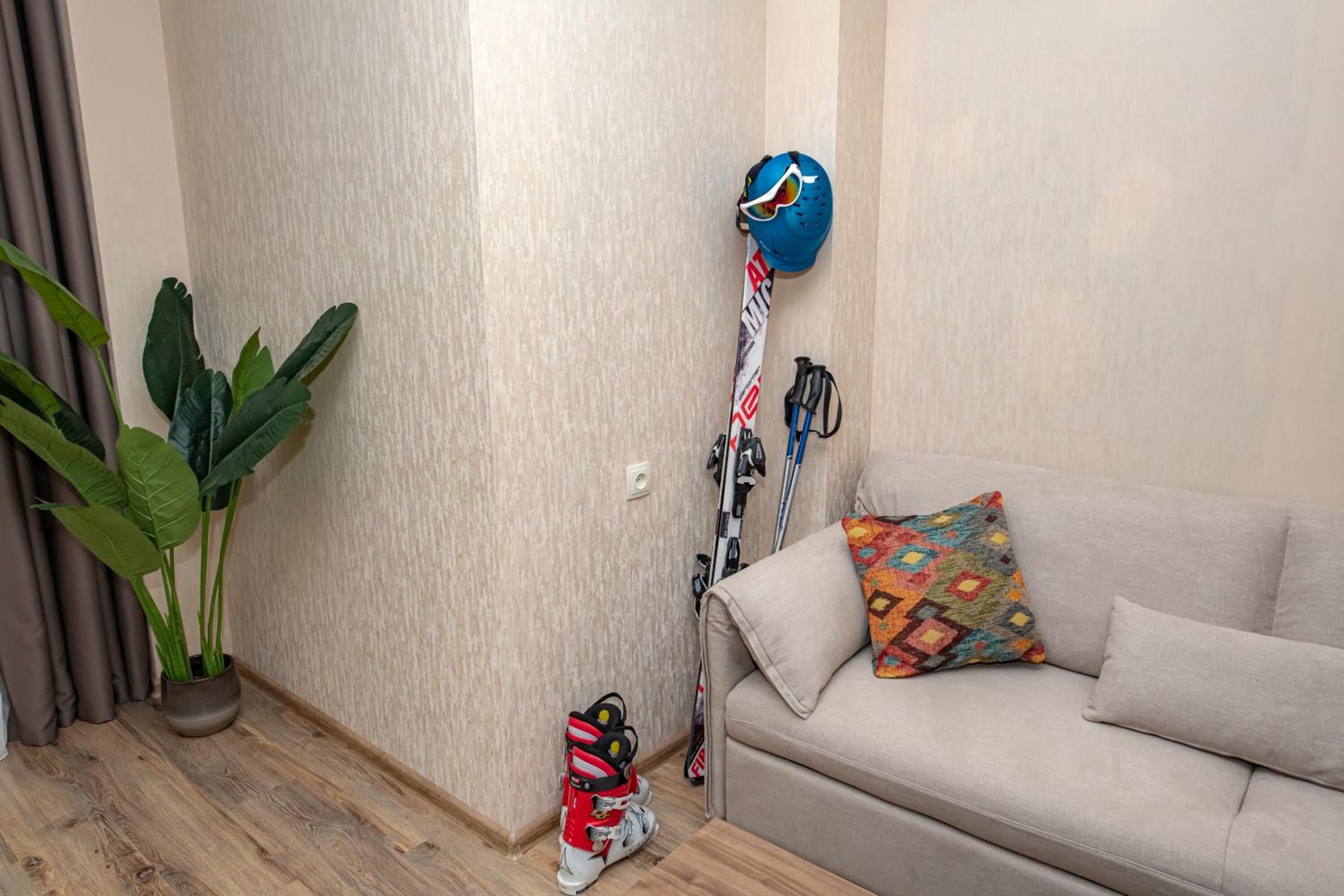 Una habitación con un sofá color beige, una almohada colorida, un casco azul, esquíes y una planta.